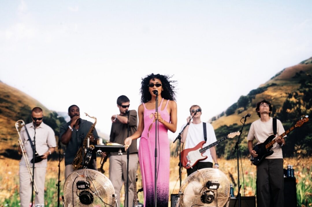 Banda a atuar ao ar livre, com uma vocalista em vestido rosa e músicos a tocar guitarra, saxofone e outros instrumentos.