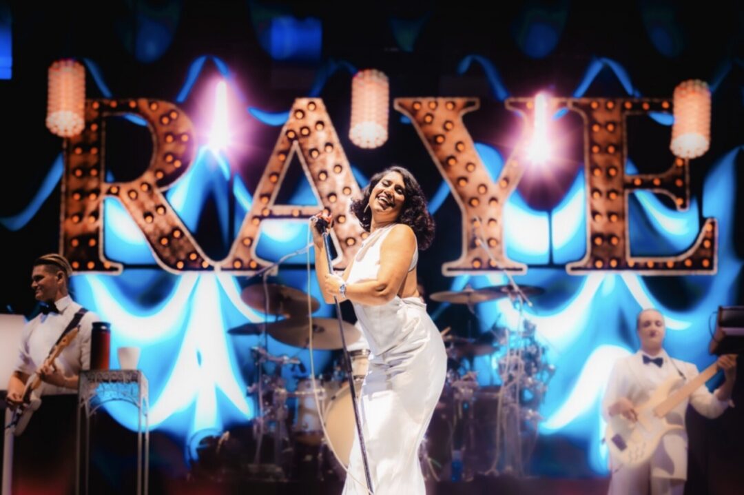 Cantora em vestido branco a sorrir no palco com a palavra “RAYE” iluminada em grande atrás, acompanhada pela sua banda.