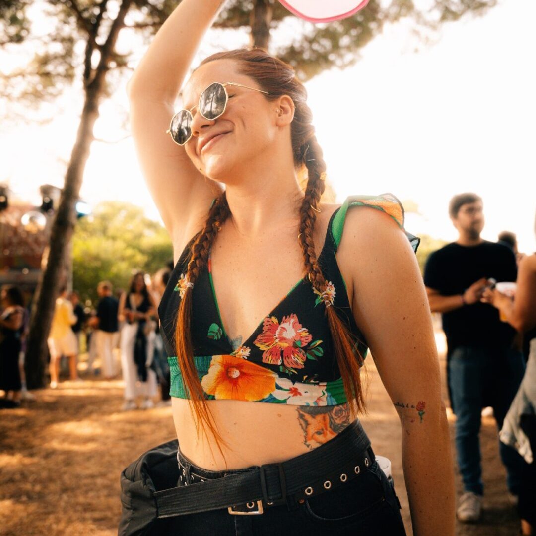 Mulher com tranças e óculos de sol a dançar ao ar livre, sorridente e com um leque na mão.