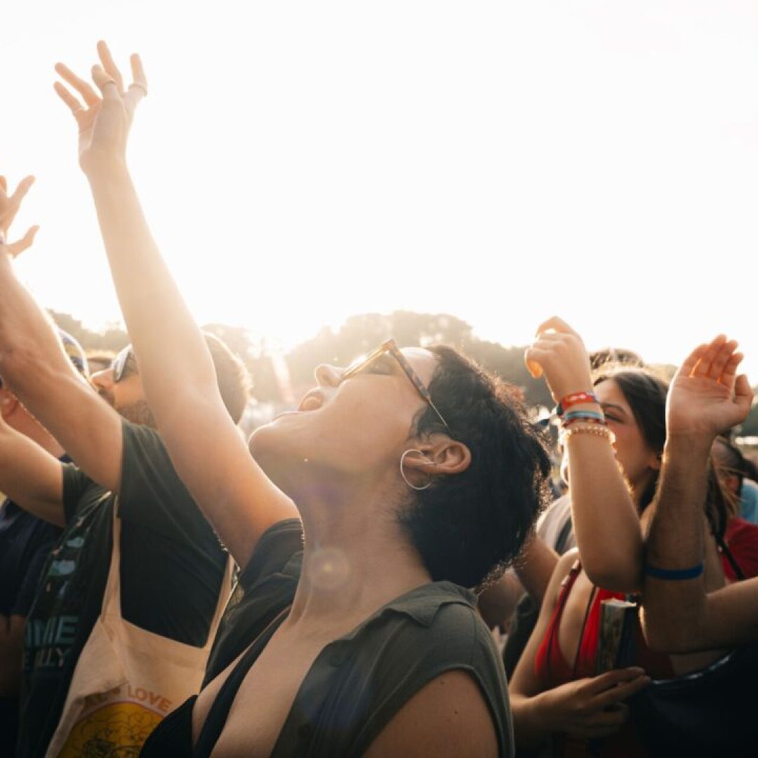 Público a dançar com os braços levantados e a cantar, iluminados pelo sol da tarde.