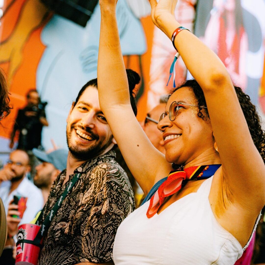 Mulher com os braços no ar a dançar e sorrir, ao lado de um homem, em frente a um palco colorido.