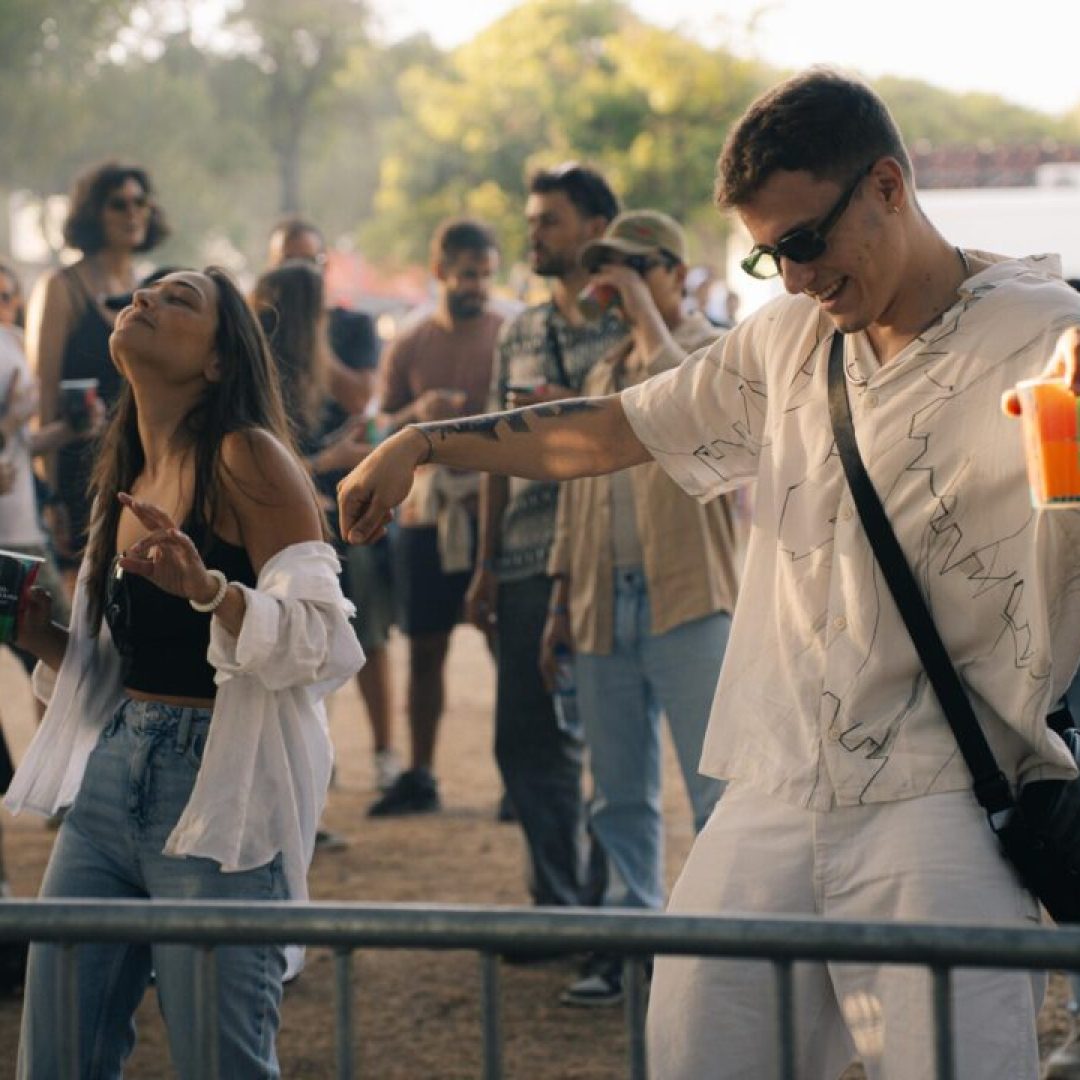 Casal jovem a dançar sorridente com copos na mão no ambiente descontraído do festival.