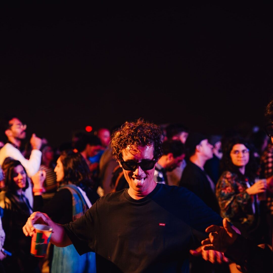 Homem a dançar animadamente com um copo na mão durante o festival MEO KALORAMA, rodeado de público.
